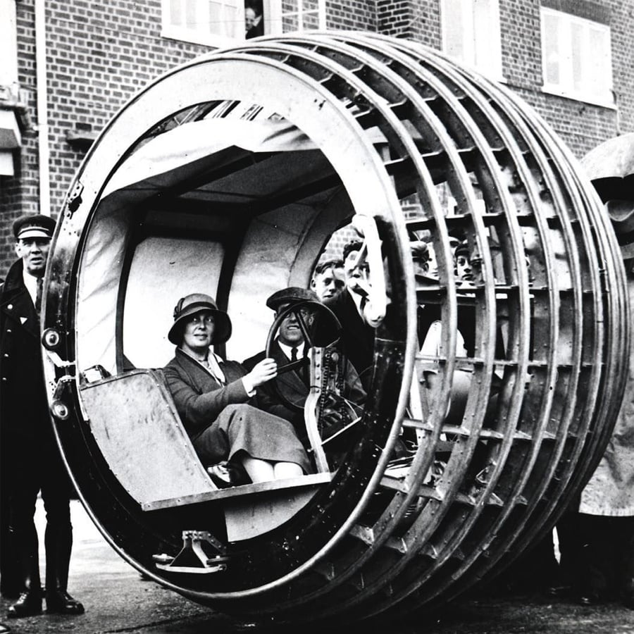 Two women sit inside a large, circular, single-wheel vehicle with an open frame, while two men stand nearby. The background shows a brick building and a person looking out of an upstairs window.