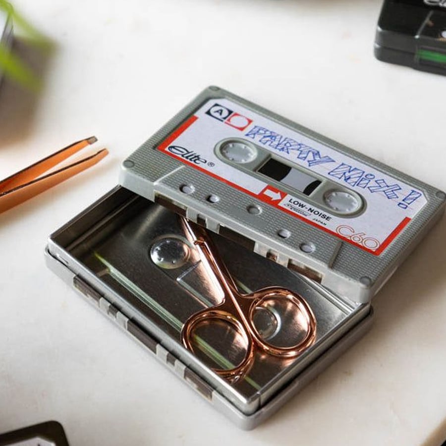 A metal case designed to look like a cassette tape sits open on a white surface, containing a pair of small scissors inside. Tweezers are nearby.