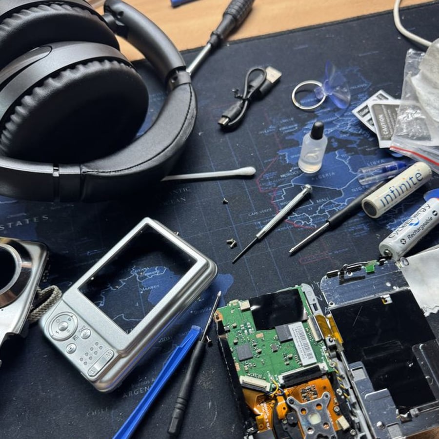A workspace with headphones, a disassembled camera, small tools, screws, a USB drive, and various electronic parts scattered on a world map desk mat.