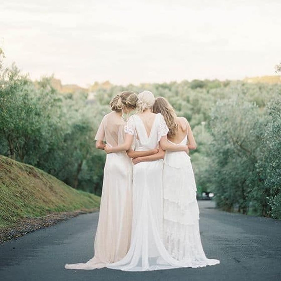 Three women in light-colored dresses stand arm in arm, facing away, on a paved road surrounded by greenery and trees under a bright sky.