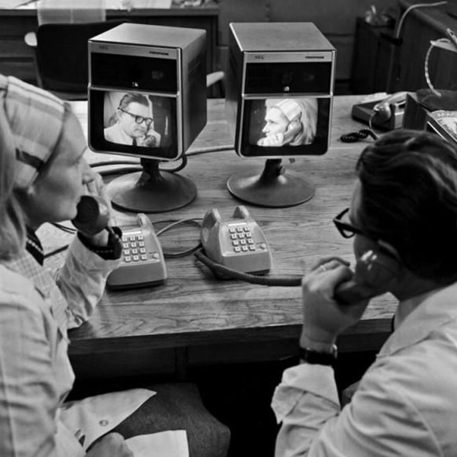 Two people use early video phones at a desk, each holding a corded handset and watching small screens showing the other person’s video image. The scene appears to be from the mid-20th century.