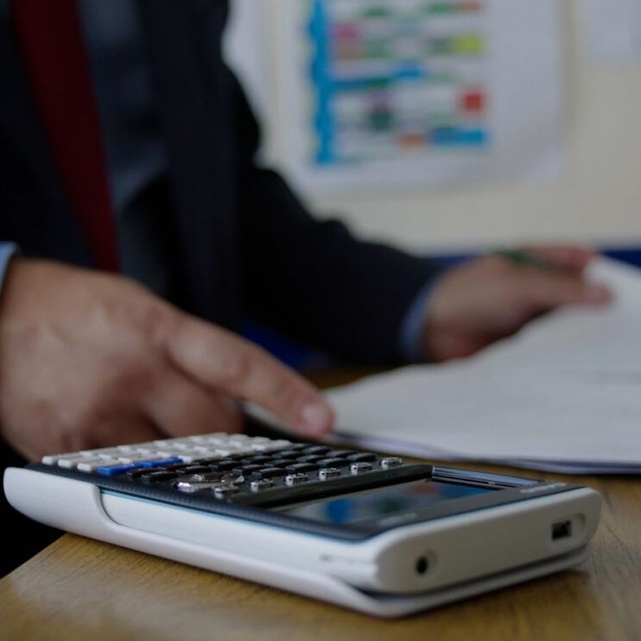 A person in a suit uses a calculator on a desk with one hand while the other hand holds an open notebook or document.