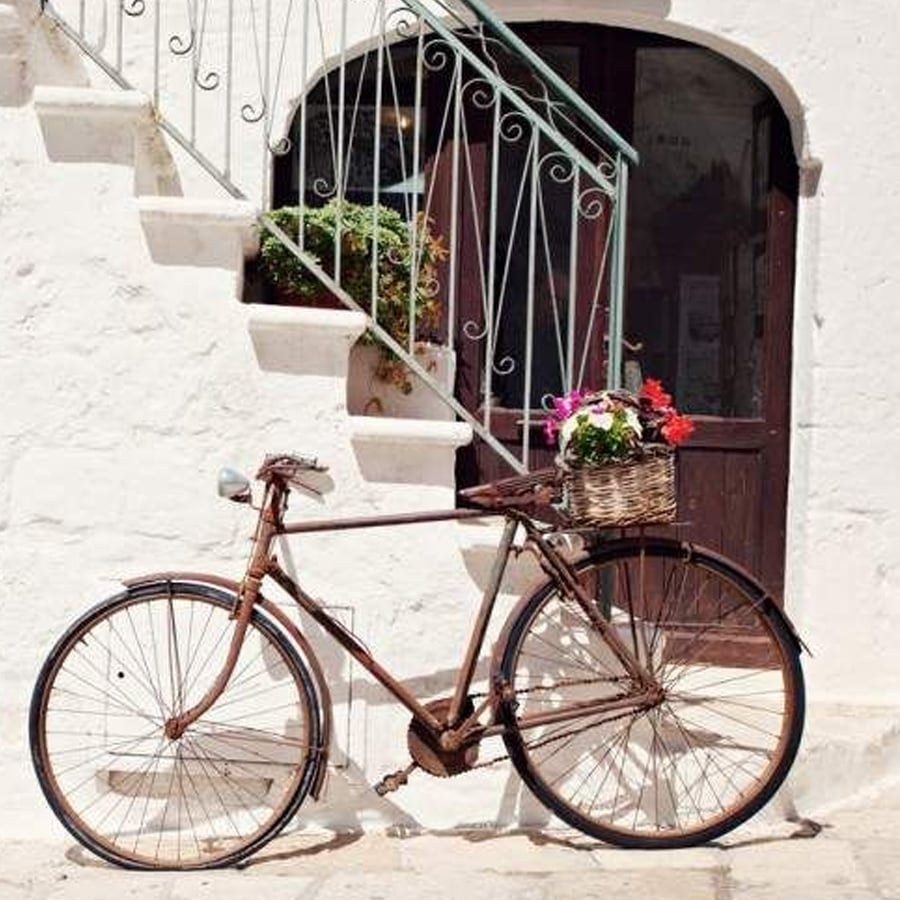 A vintage bicycle with a basket of colorful flowers leans against a white stone wall beneath a staircase with green railings.