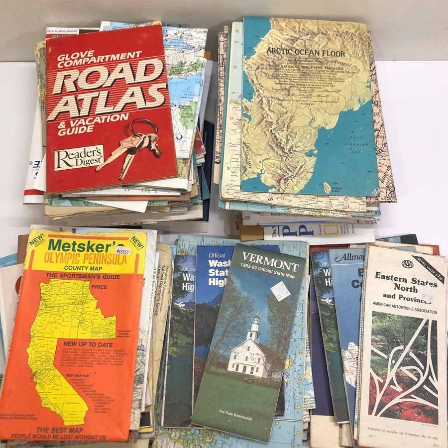 Stacks of vintage road atlases, maps, and guidebooks are spread out on a white surface, showing covers with colorful titles, maps, and illustrations of various U.S. regions and landmarks.