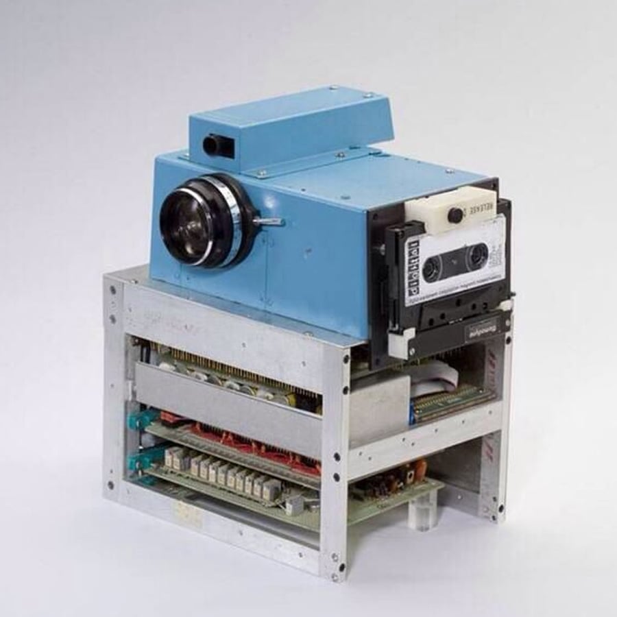 A bulky, vintage digital camera with a blue body and visible internal circuitry, sitting on a metal frame with circuit boards and electronic components exposed.
