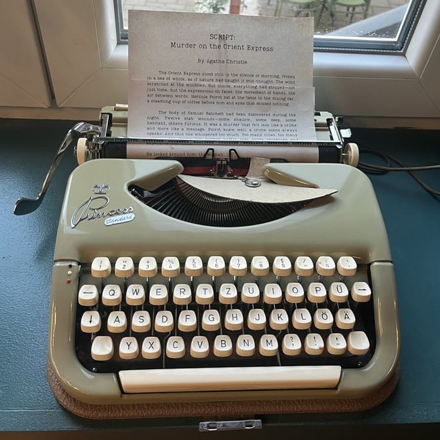 A vintage Princess typewriter sits on a desk by a window, with a script titled “Murder on the Orient Express” by Agatha Christie loaded in the roller.
