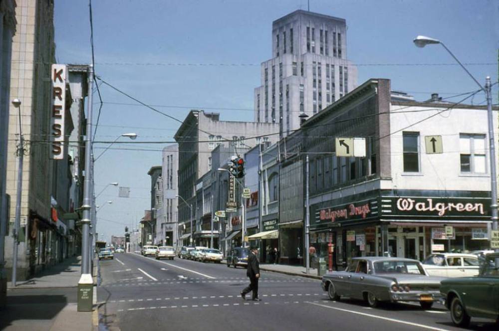 A man crosses a city street lined with vintage cars and storefronts, including Kress, Walgreen, and Walgreen Drugs. Tall buildings and a traffic light are visible under a clear blue sky.