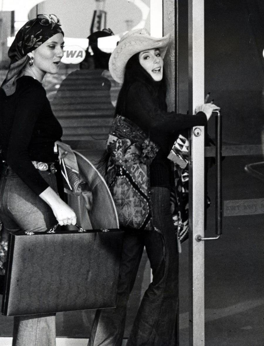 Two women in 1970s outfits, one wearing a headscarf and holding a briefcase, the other in a cowboy hat and patterned jacket, are entering a glass door, smiling at the camera.