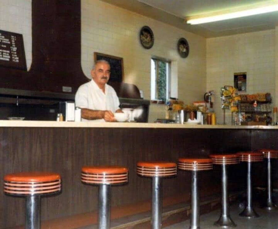 A man in a white shirt stands behind a classic diner counter with red stools. The counter displays condiments, napkins, and dispensers, with menus and decorations on the wall behind him.