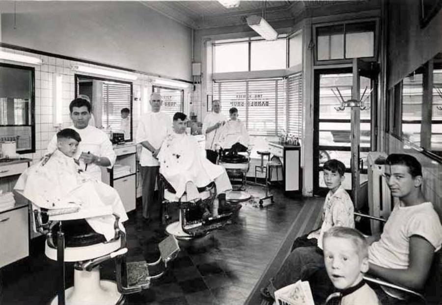 Black-and-white photo of a vintage barbershop with barbers cutting hair and several boys waiting. Large mirrors and windows line the walls; the atmosphere is busy and casual.