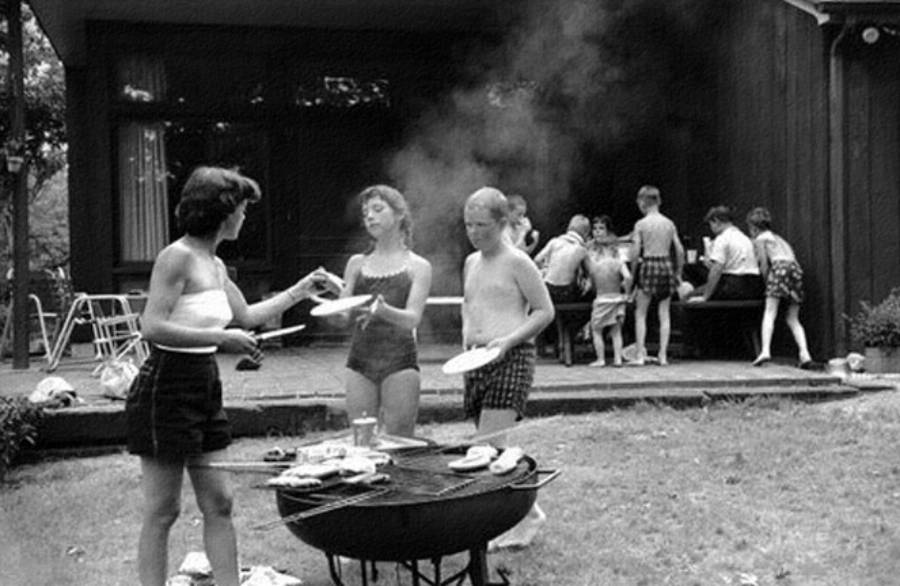 A group of people in swimsuits gather for a backyard barbecue. Three stand by a grill with food, while others eat and talk on a patio in the background. Smoke rises from the grill, and the scene is lively and summery.