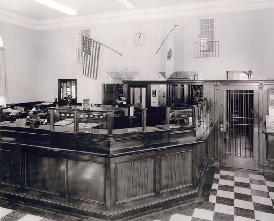 Black and white photo of an old bank interior with wooden counters, an American flag, a vault door, desks, and a clock on the wall. The floor has a checkered tile pattern.