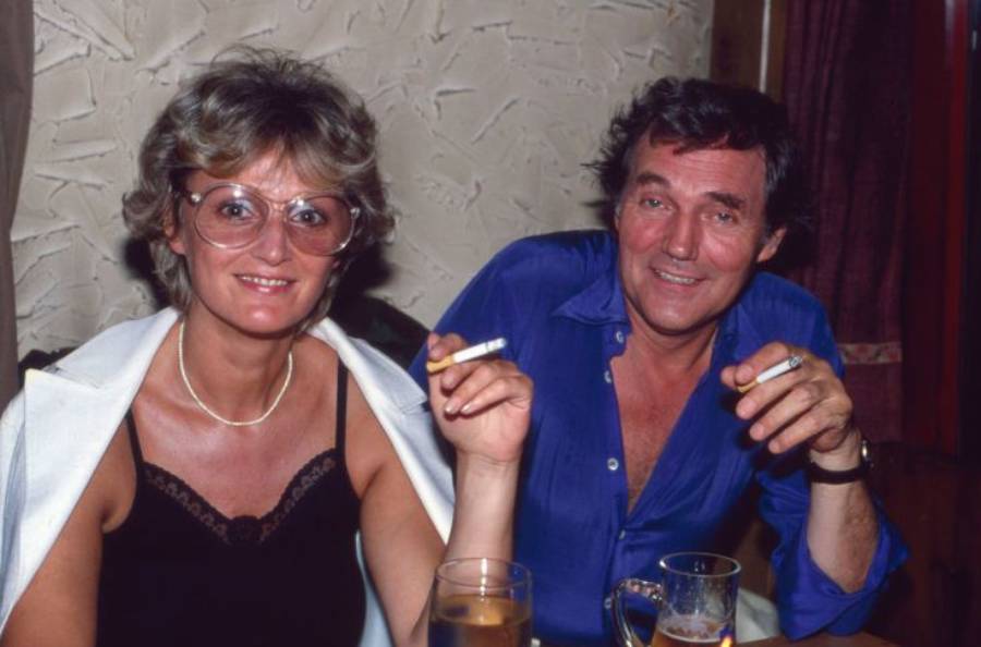 A smiling woman and man sit together at a table, each holding a cigarette. The woman wears glasses and a black top, while the man wears a blue shirt. They both have drinks in front of them.