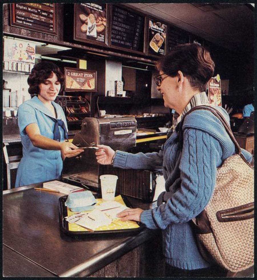 A woman in a blue sweater hands money to a smiling fast-food worker behind the counter. A tray with food, drinks, and wrappers sits on the counter. The menu is visible in the background.