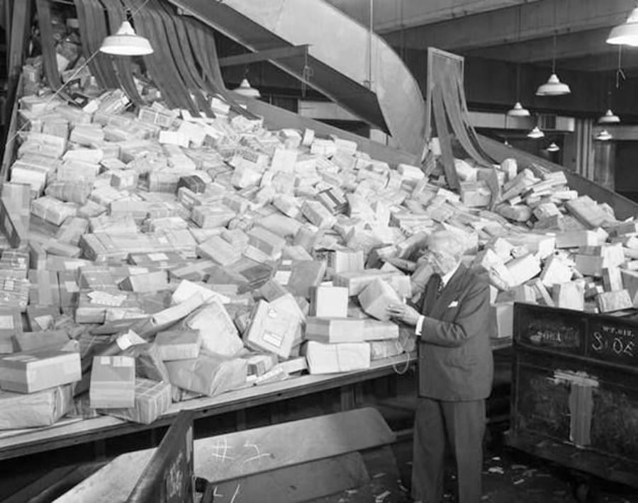 A man in a suit stands beside a massive pile of mail parcels on a sorting conveyor inside a large warehouse, under hanging lights.