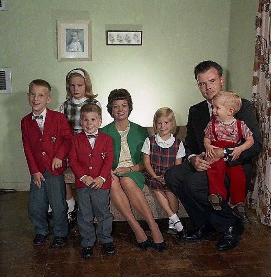 A family of six poses indoors. Two adults sit on a couch, with a young girl beside them. Three children, two boys in red jackets and a girl in plaid, stand nearby. The group smiles in a warmly lit room.