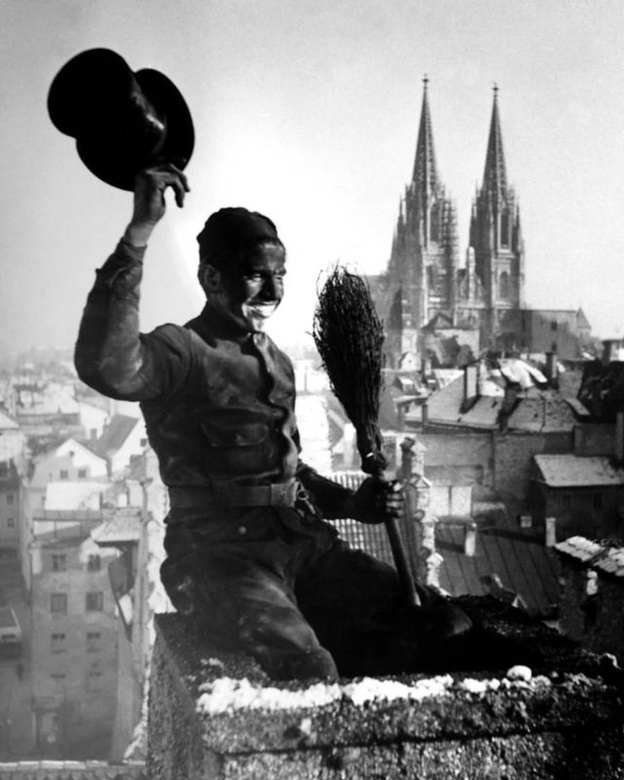 A smiling chimney sweep, face and clothing covered in soot, sits on a rooftop chimney holding a broom and tipping his hat. A cityscape and large Gothic cathedral towers are visible in the background.