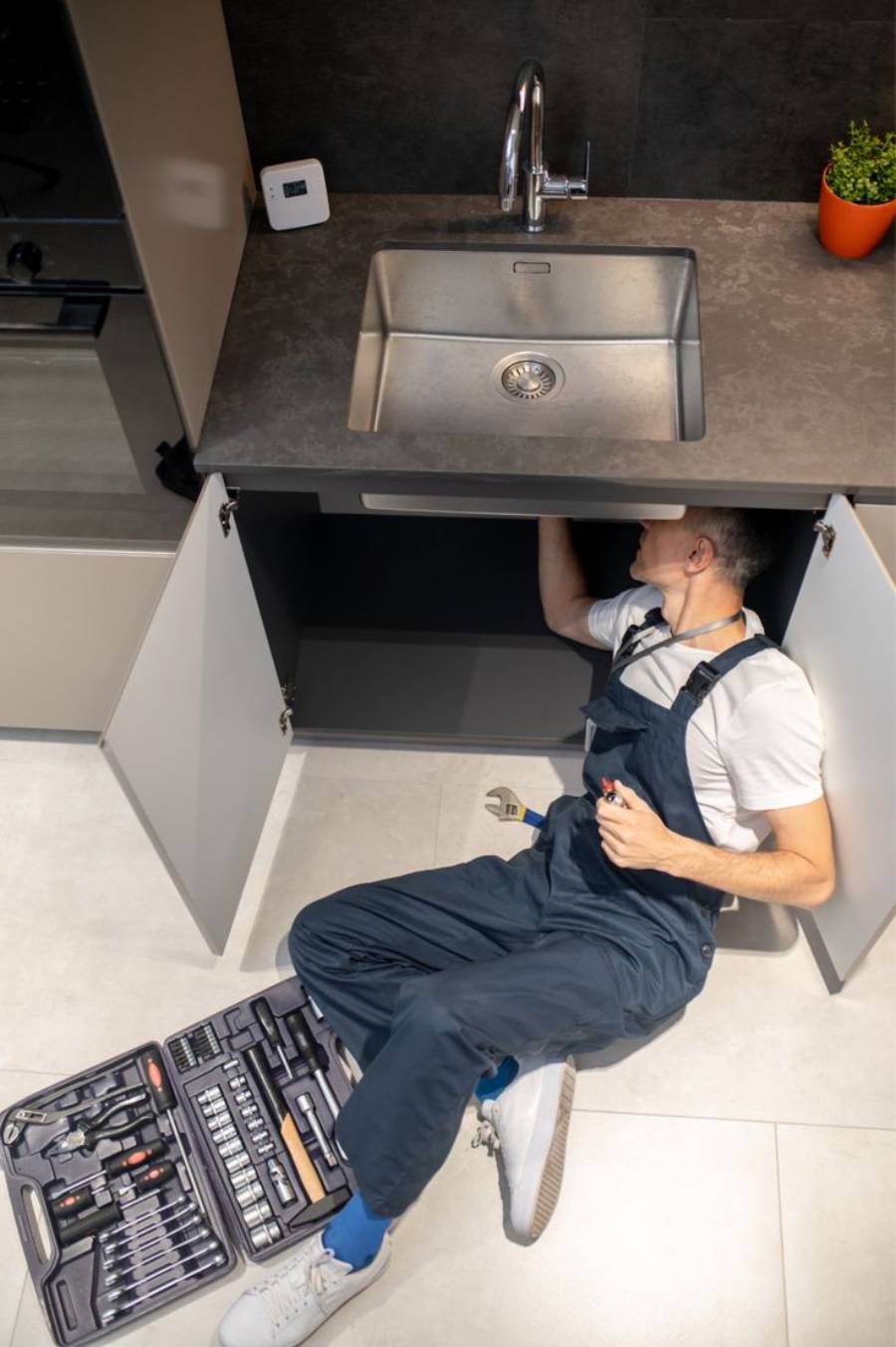 A plumber in overalls lies on the floor, working under a kitchen sink with tools spread out nearby. The sink's cabinet doors are open, and a small orange plant pot sits on the countertop.