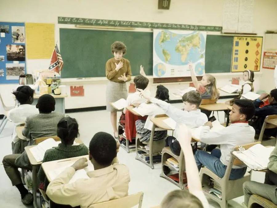 A teacher stands at the front of a classroom, addressing students with raised hands. World maps, charts, and posters decorate the classroom walls. Students sit at desks, appearing engaged in the lesson.