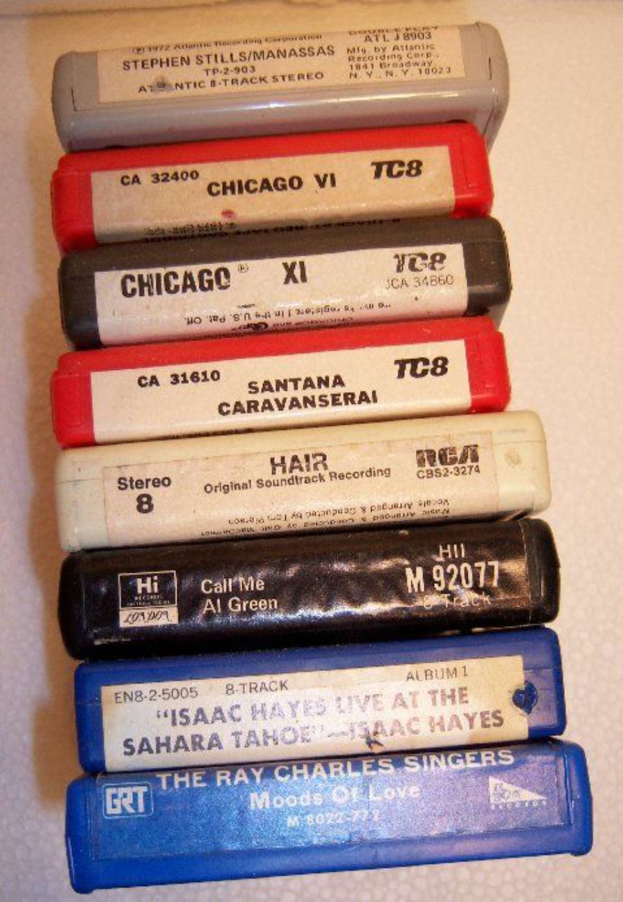 A stack of eight vintage 8-track tapes is shown, featuring albums by artists like Chicago, Santana, Al Green, Isaac Hayes, and the Ray Charles Singers. The tapes are various colors and have worn labels.