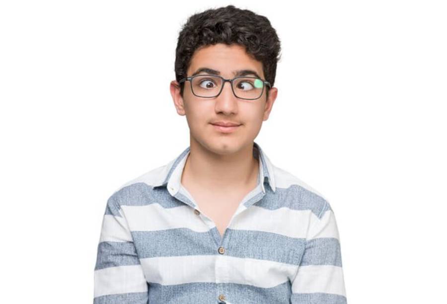 A young person with curly hair and glasses is crossing their eyes, wearing a blue and white striped shirt and standing against a plain white background.
