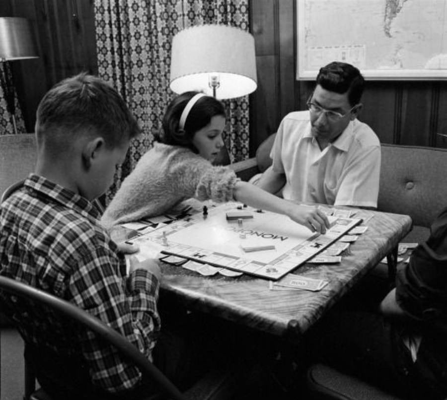 A family of three sits around a small table playing Monopoly in a cozy, wood-paneled room. The girl reaches across the board while the boy looks at his cards and the man watches.