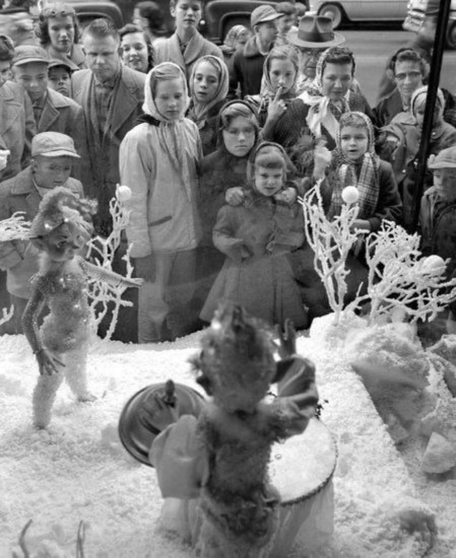 A crowd of children and adults gaze through a store window at a snowy holiday display featuring small costumed figures and white trees, with vintage cars visible in the background.