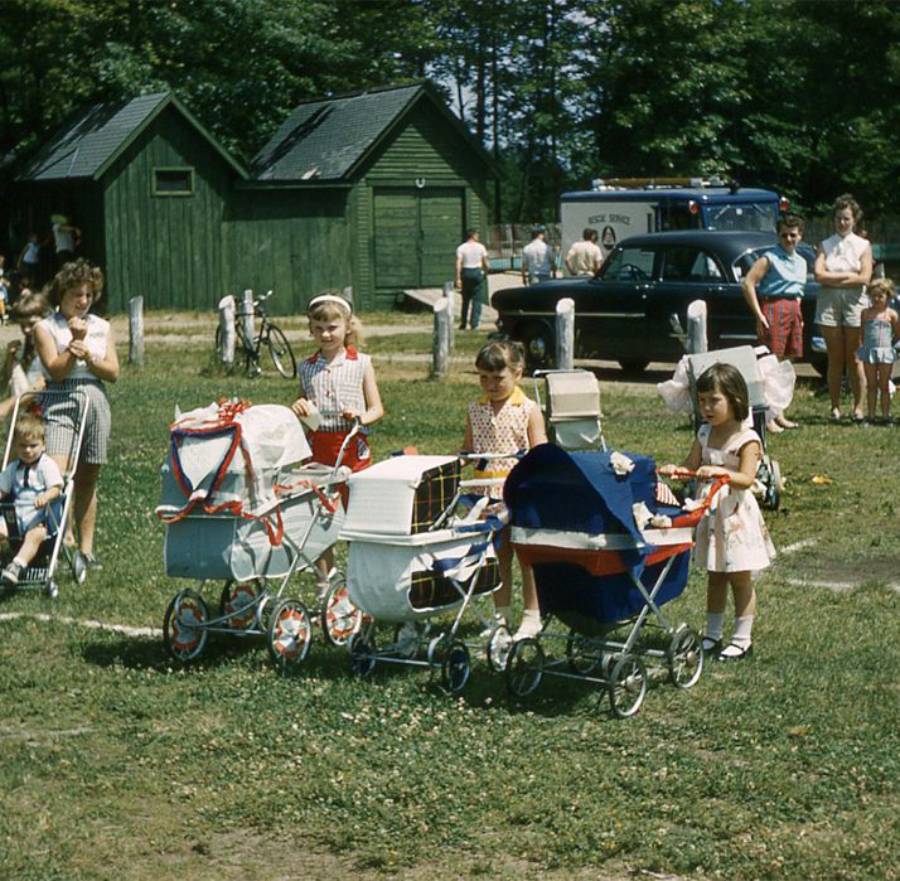 Four young girls stand outdoors with colorful toy prams, while adults and children watch in the background. There are green sheds, trees, and a black vintage car in the scene, suggesting a park or community event.