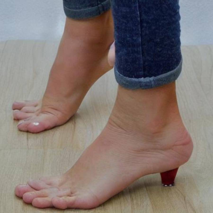 A person standing on a wooden floor with shoes designed to look like bare feet, with a small red high heel attached under one heel.