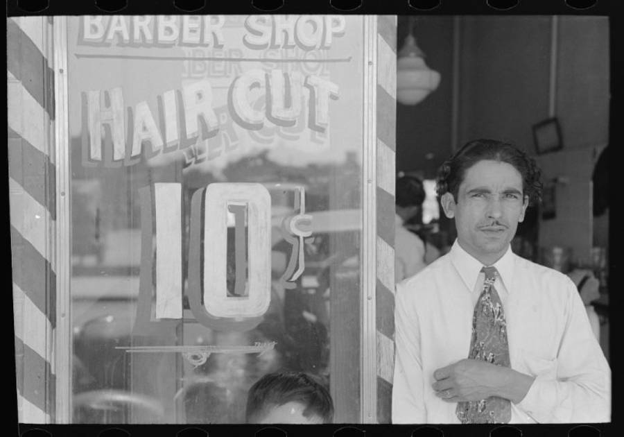 A man in a white shirt and tie stands next to a barber shop window advertising haircuts for 10 cents. The shop interior and part of a child’s head are visible in the background.