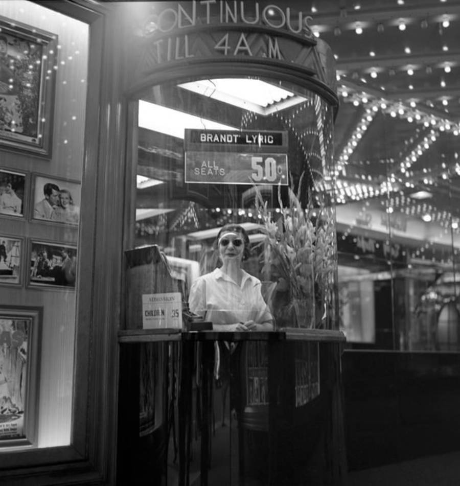 A ticket booth attendant in sunglasses sits behind glass at a vintage theater, with movie posters, bright lights, and a sign advertising "All seats 50¢" and "Continuous till 4 A.M." visible.