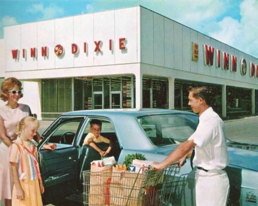 A family loads groceries into their car outside a Winn-Dixie supermarket on a sunny day. A man pushes a cart full of groceries, while a woman and two children stand nearby, all smiling.
