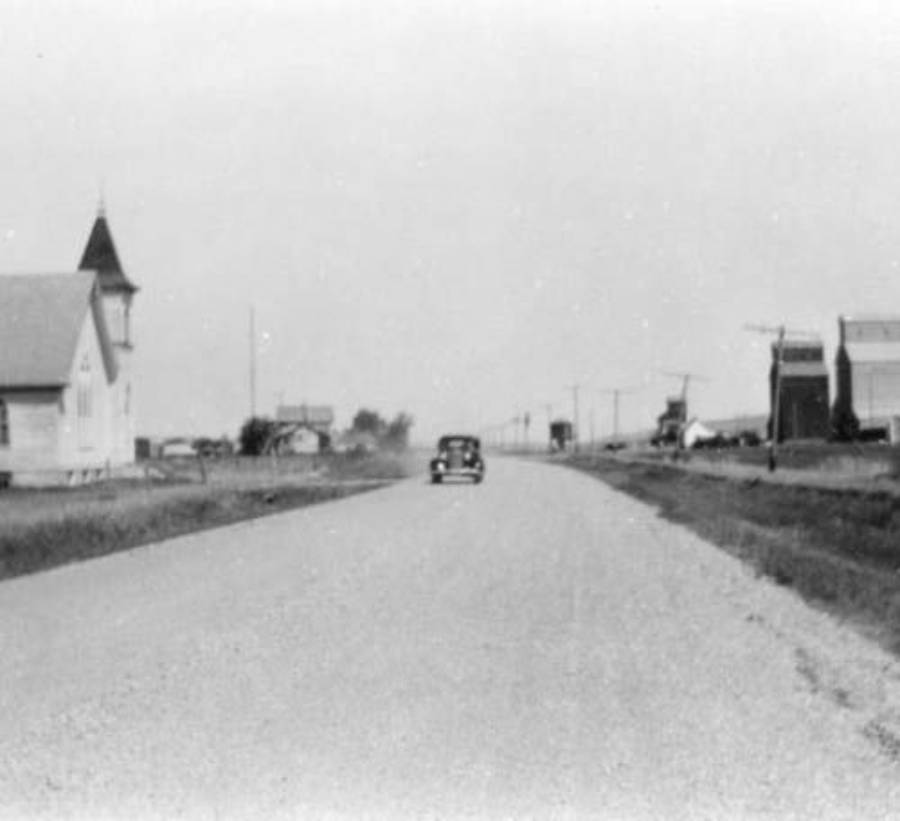 A black-and-white photo of a vintage car driving down a wide, empty rural road, with a church on the left and a few buildings and power lines along the roadside in the distance.