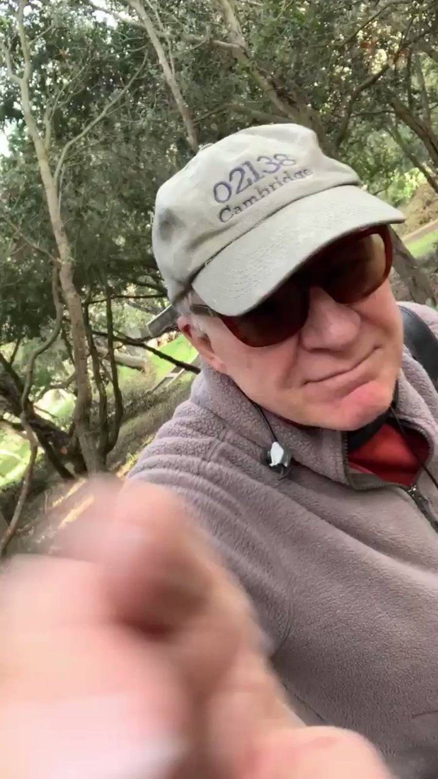 An older man wearing a "02138 Cambridge" cap, sunglasses, and a fleece jacket stands outdoors in a wooded area, reaching toward the camera with one hand.