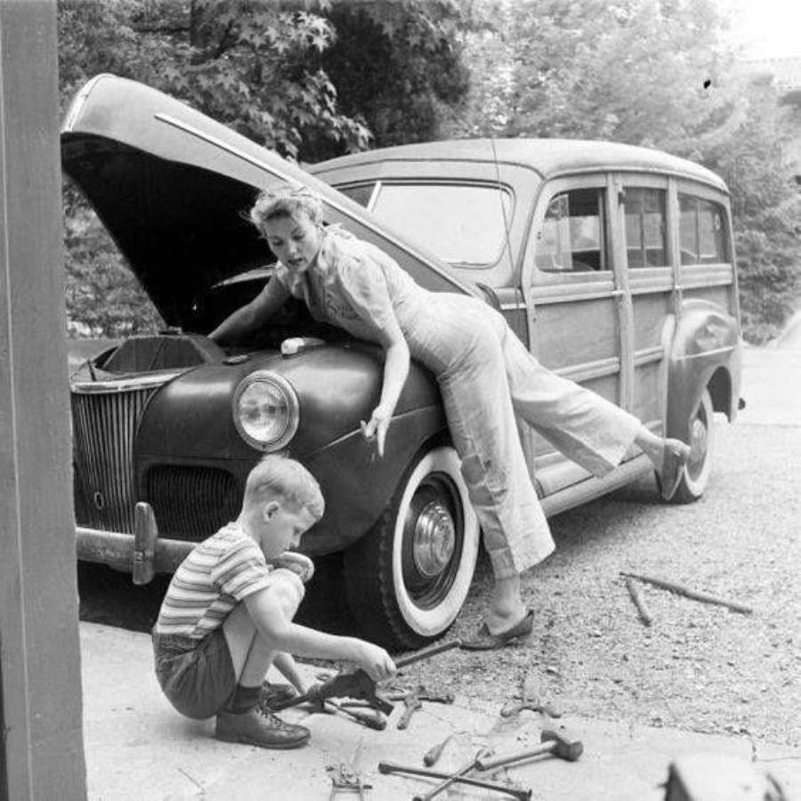 A woman leans over the engine of a vintage car with the hood open, while a young boy sits on the ground nearby, sorting through tools scattered around him. Trees and bushes are visible in the background.