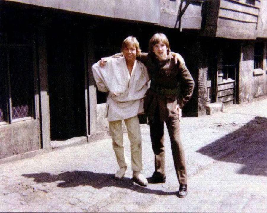 Two young men stand outside on a cobblestone street, smiling with arms around each other. One wears light-colored clothes, the other wears a brown outfit. The old buildings behind them are weathered and shadowed.