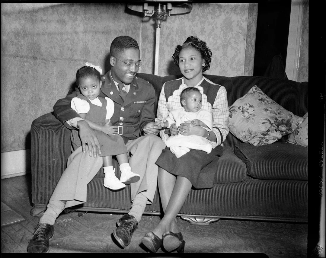 A smiling Black family sits on a couch: a man in a military uniform holds a young girl, while a woman holds a baby. The room appears warm and cozy, with patterned wallpaper and a pillow on the sofa.