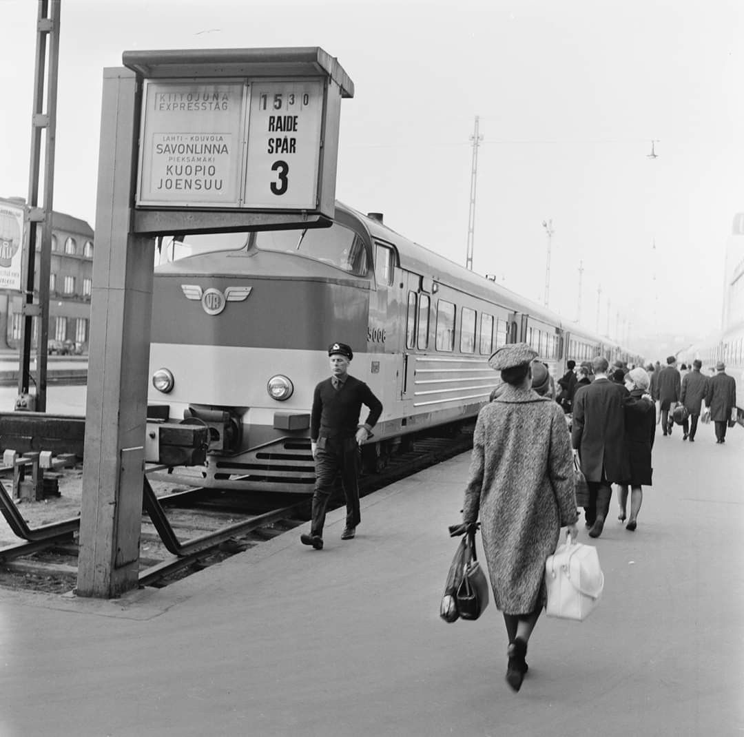 Black and white photo of people walking on a train platform. A train is stopped, with a conductor nearby. A sign shows destinations: Savonlinna, Kuopio, Joensuu. People carry bags and wear coats and hats.