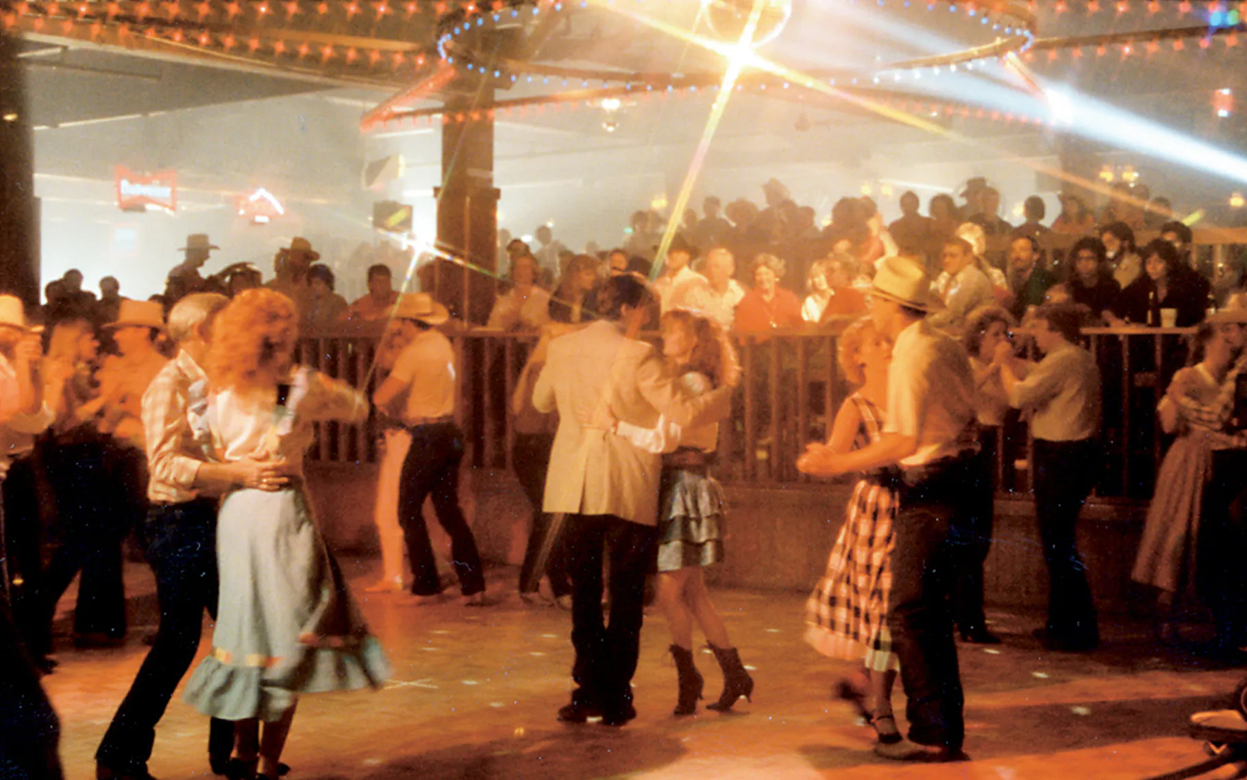 People in Western attire dance under colorful lights in a crowded nightclub, while a large audience watches from behind a wooden railing. The scene is lively and filled with country-themed decorations.