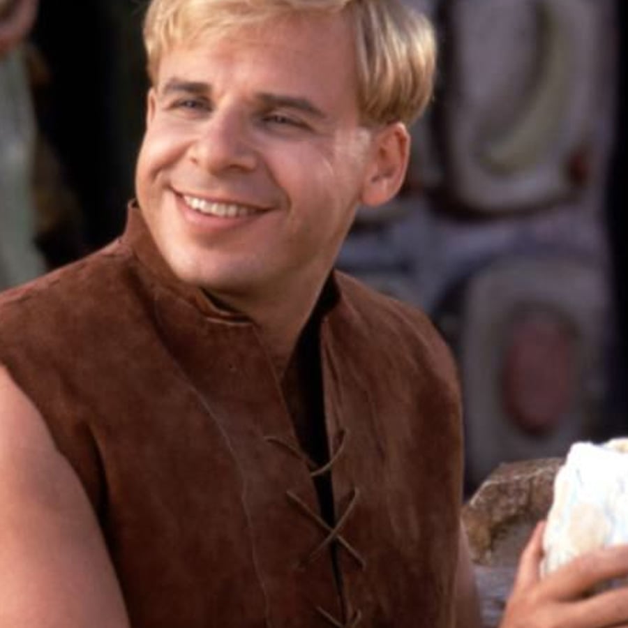 A man with short blond hair smiles while wearing a brown sleeveless vest. He is outdoors, and the background is slightly blurred with stone and greenery visible.