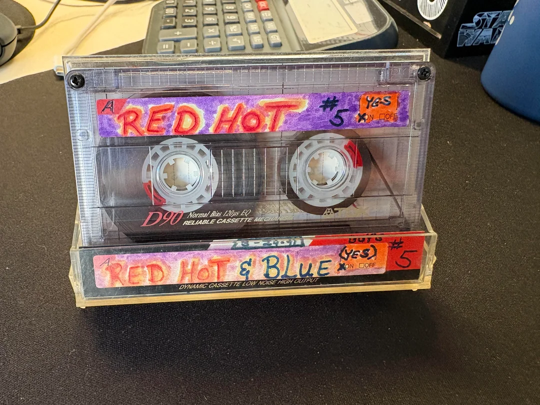 A labeled cassette tape reads "RED HOT" on side A and "RED HOT & BLUE" on side B, with handwritten notes, the number 5, and "YES! XN COFF," placed on a desk near a calculator.