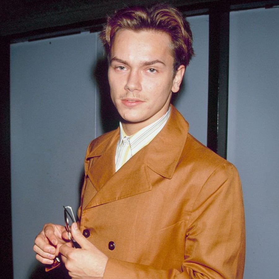 A young man wearing a brown double-breasted coat stands indoors, holding sunglasses in his hand and looking directly at the camera with a calm expression.