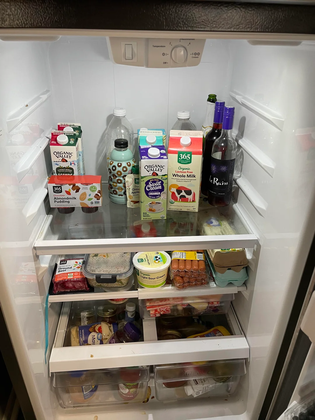 An open refrigerator contains milk, juice, eggs, berries, bottles of wine, yogurt, butter, various packaged foods, and fresh produce in drawers. Shelves are illuminated by the fridge light.