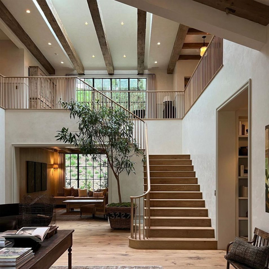 Spacious modern living room with wooden staircase, large windows, ceiling beams, indoor tree, and cozy seating area with warm lighting. Neutral tones and natural light create an inviting, open atmosphere.