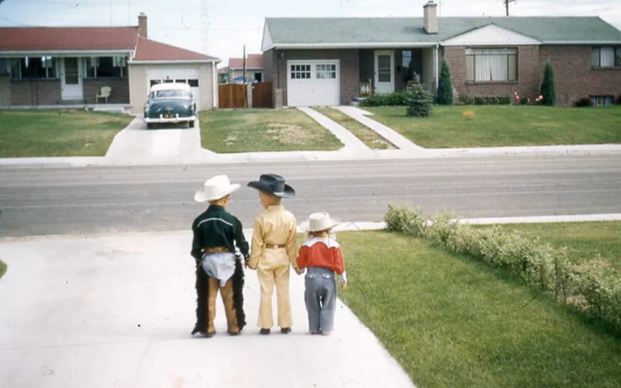 Three children in cowboy costumes stand on a driveway, facing a suburban street with single-story houses and a vintage car parked in a garage across the road.
