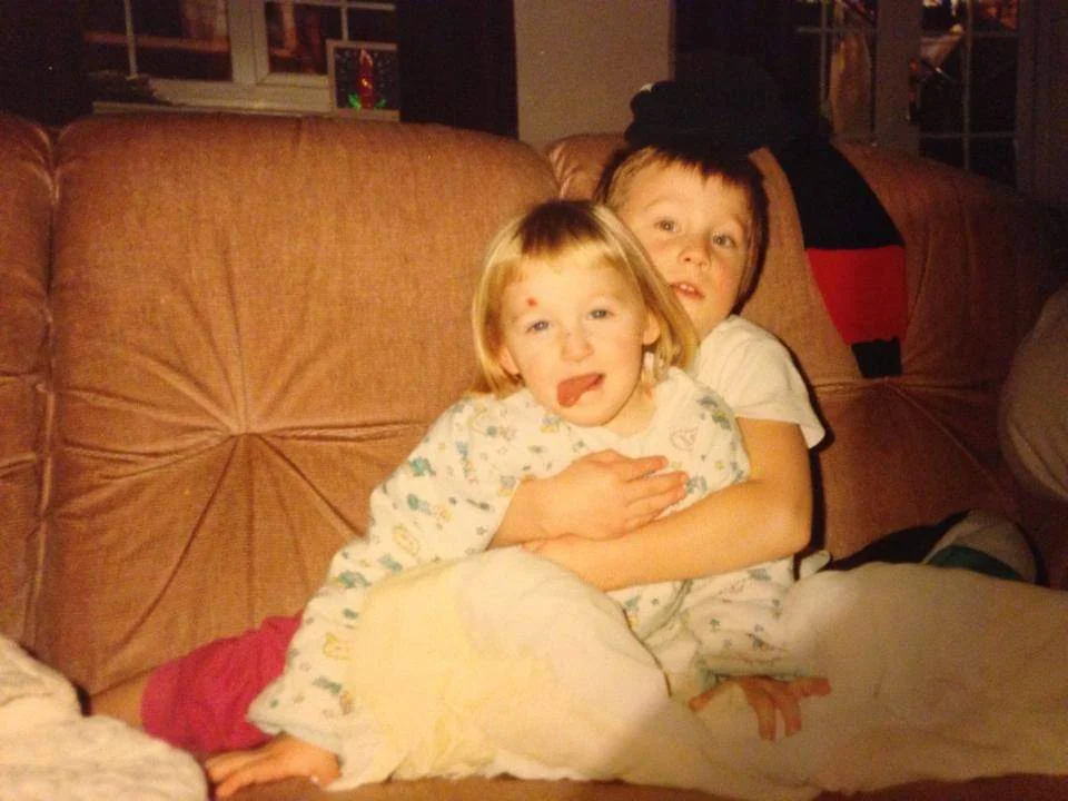 Two young children sit on a brown couch. The older child hugs the younger one from behind. The younger child sticks their tongue out playfully. Both are in pajamas and covered with a blanket.