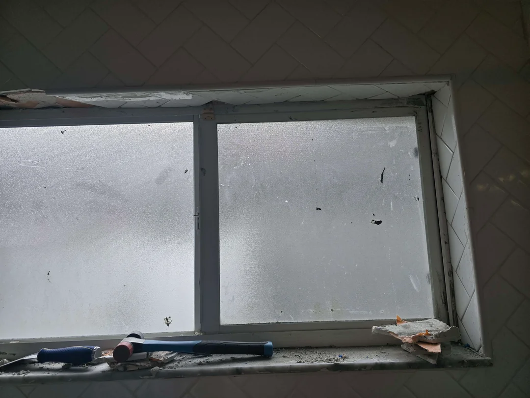 A dirty, fogged-up window with debris and tools on the sill, including a hammer, screwdriver, and some papers. The tile wall around the window appears to be chipped and in need of repair.