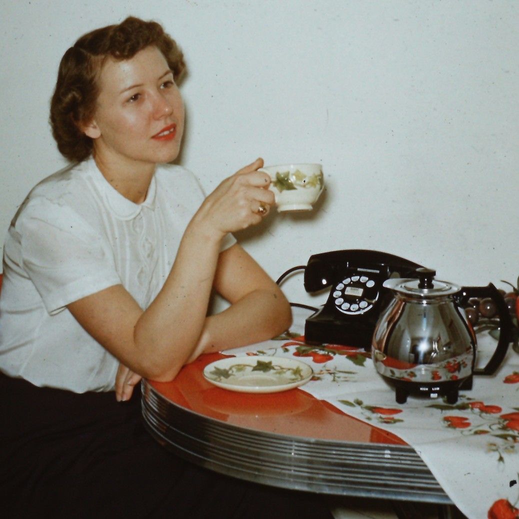 A woman in a white blouse sits at a retro table, holding a teacup. A plate, teapot, rotary phone, and floral tablecloth are also on the table. She looks thoughtful.