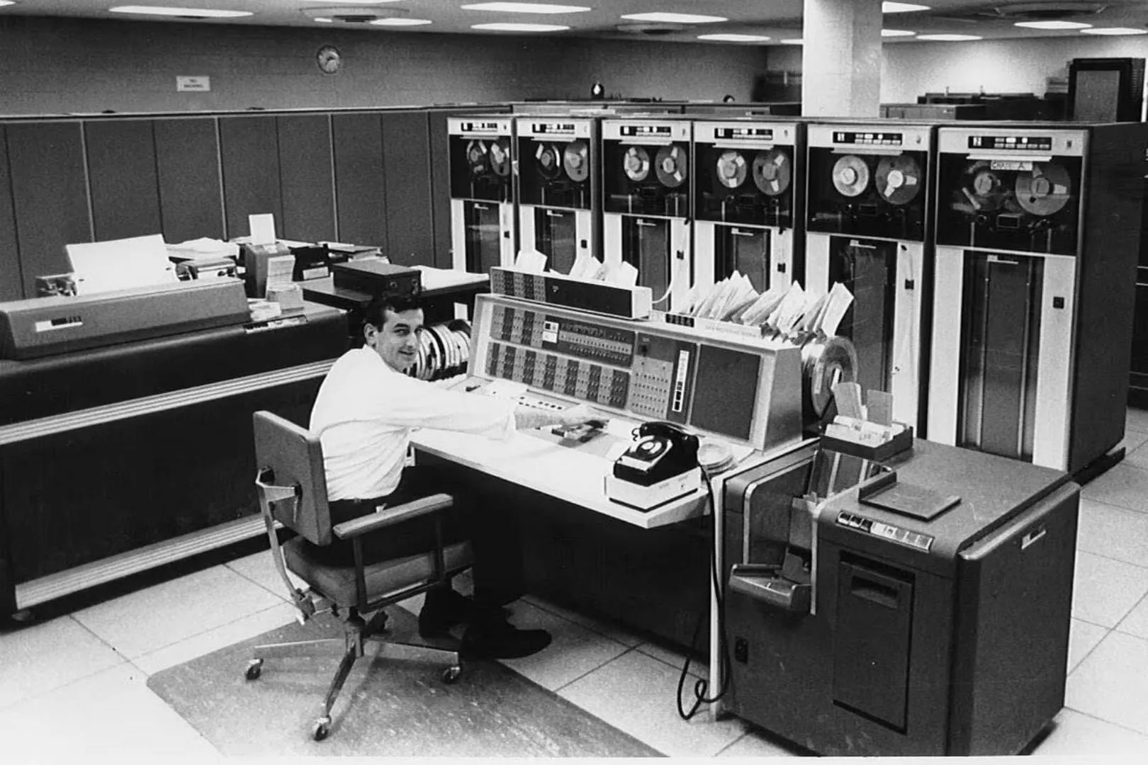 A man sits at a vintage computer console surrounded by large tape reel machines and office equipment in a retro data center room.