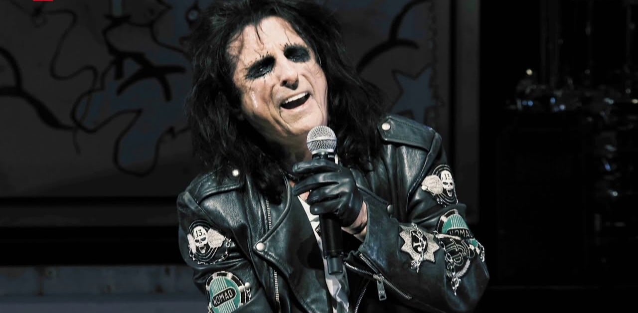 A person with long dark hair, dramatic black eye makeup, and a black leather jacket sings passionately into a microphone while wearing black gloves.